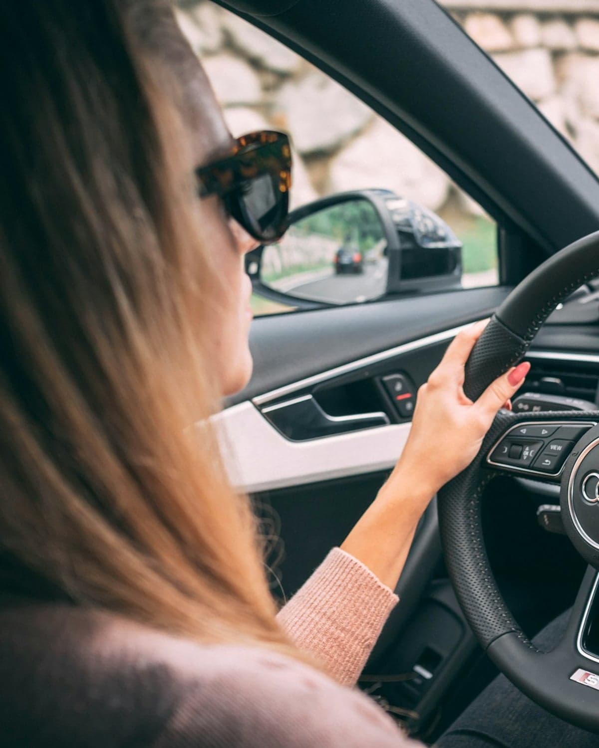 A driver with both hands on the steering wheel, the road behind visible in the side mirror