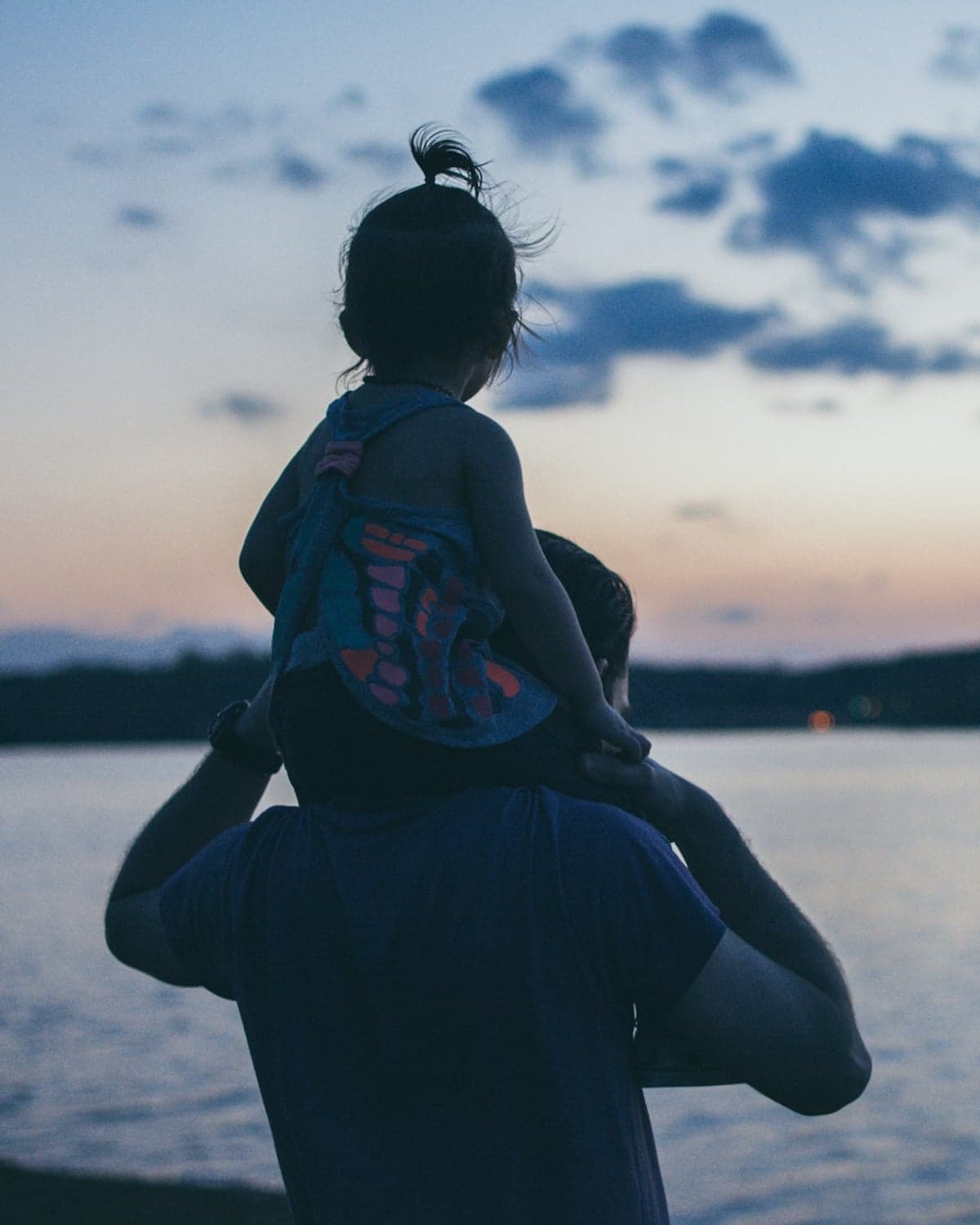 A parent carrying a young child on their shoulders beside the water at dusk